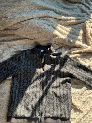 Svart ziptröja från Lacoste - Snygg mörkgrå ziptröja från Lacoste med diskret geometriskt mönster och klassisk krokodillogga på bröstet. Tröjan har hög krage, hel dragkedja och ribbade muddar. Perfekt för en stilren och avslappnad look.