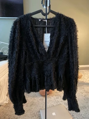 Svart fluffig omlottopp från NA-KD - Säljer en svart fluffig omlottopp från NA-KD med v-ringning och långa ärmar. Toppen har en unik textur och knyt i midjan som ger en snygg siluett. Aldrig använd, med prislapp.