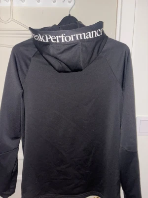 Svart Peak Performance hoodie - Säljer en svart hoodie från Peak Performance med dragkedja framtill och huva. Loggan syns på bröstet och texten 'Peak Performance' står tryckt på huvan. Hoodien har långa ärmar och två fickor med dragkedja. Materialet känns sportigt och stretchigt.