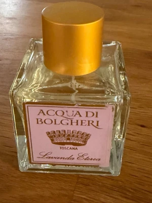 Acqua di Bolgheri Lavanda Eterea parfym - En elegant parfym i en fyrkantig glasflaska med guldig kork. Doften är Lavanda Eterea från Acqua di Bolgheri, Toscana. Flaskan har en ljusrosa etikett med guldiga detaljer och rymmer en fräsch och blommig doftupplevelse.
