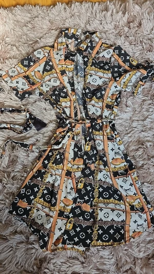 Mönstrad kortklänning med bälte - Snygg kortklänning med svart, vit och beige mönster, detaljer i guld och orange samt bälte i midjan. Klänningen har korta ärmar, krage och knappar framtill och fickor. Perfekt för dig som gillar statement-plagg med coola kedje- och bältesdetaljer.