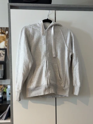 Ljusgrå zip hoodie Russedress - Ljusgrå hoodie med dragkedja från Russedress. Klassisk modell med huva och vita dragsnören, samt två stora fickor framtill. Tillverkad i mjukt sweatshirtmaterial, perfekt för chill dagar. Ribbstickade muddar vid ärmslut och nederkant.