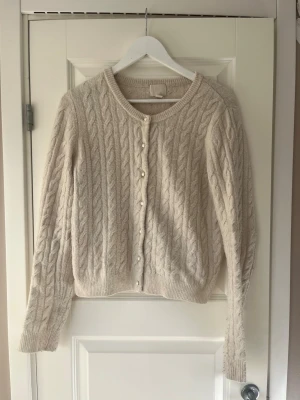 Kabelstickad kofta - Säljer en vit/beige kabelstickad kofta med rund halsringning och pärlknappar framtill. Koftan har långa ärmar och ribbade muddar vid ärmslut och nederkant. Riktigt mysigt material och perfekt för en kylig dag. Har varit en av mina favorit tröjor och är väl använd men säljer då den tyvärr blivit för kort för mig💗