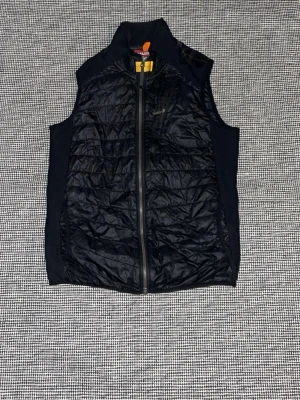 Svart dunväst från Parajumpers - ✅priset är inte hugget i sten✅Snygg svart dunväst från Parajumpers med hög krage och dragkedja framtill. Västen har quiltad design och är tillverkad i ett glansigt material som ger en sportig look. Perfekt att slänga över en hoodie eller tröja för extra värme.