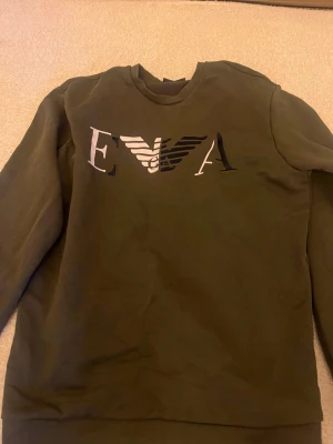 Olivgrön sweatshirt från EA7 - Säljer en olivgrön sweatshirt från EA7 med stort logotryck på bröstet. Tröjan har rund halsringning och långa ärmar. Tillverkad i mjukt material som känns skönt mot huden. Perfekt för en avslappnad och stilren look.