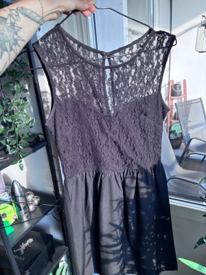 Svart spetsklänning med ärmlös design - Snygg svart klänning med spets upptill och ärmlös design. Överdelen har transparent spets med blommönster och rund halsringning, medan kjolen är enkel och slät. Perfekt för dig som gillar en elegant och stilren look.