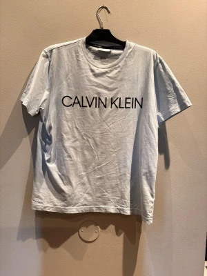 Vit t-shirt från Calvin Klein - Snygg vit t-shirt från Calvin Klein med stor svart logga över bröstet. Klassisk rund halsringning och korta ärmar. Tillverkad i mjuk bomull för en skön känsla och enkel stil.