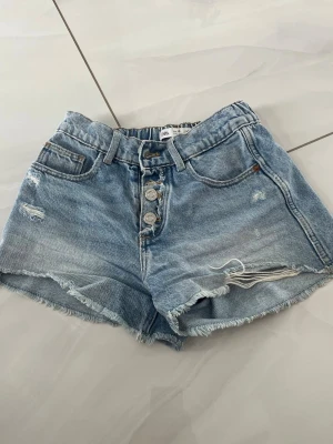 Ljusblå denimshorts med knappar - Säljer ett par ljusblå denimshorts med fransig kant och slitna detaljer. Shortsen har hög midja, fyra synliga knappar framtill och klassiska fem fickor. Perfekta för varma dagar och en avslappnad stil.