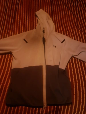 Tvåfärgad hoodie från Under Armour - Snygg hoodie från Under Armour i två färger, ljusgrå upptill och mörkgrå nedtill. Dragkedja framtill, huva och diskret logga på bröstet. Perfekt för chill eller träning. Mjukt material och skön passform.