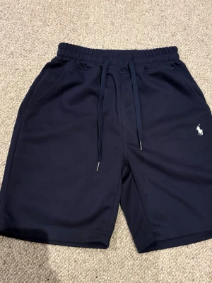 Marinblå shorts från Polo Ralph Lauren - Snygga marinblå shorts från Polo Ralph Lauren med dragsko i midjan och klassisk logga broderad i vitt på benet. Shortsen har sidofickor och är tillverkade i mjuk bomull, perfekta för en avslappnad stil.