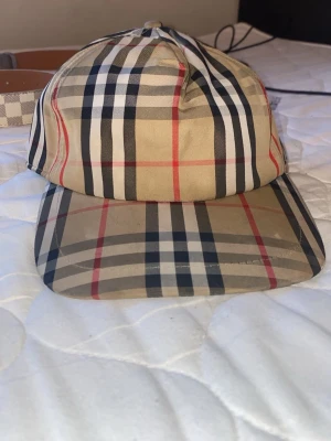 Burberry beige rutig keps - Säljer en klassisk Burberry keps i beige med det ikoniska rutmönstret i svart, vitt och rött. Kepsen har böjd skärm och justerbar snapback baktill. Märkt med 'BURBERRY LONDON ENGLAND' på baksidan. Perfekt för dig som gillar streetstyle och designeraccessoarer.