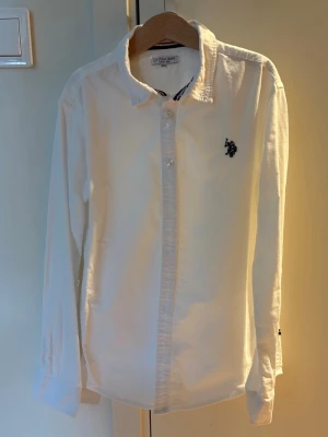 Vit långärmad skjorta från U.S. Polo Assn. - Klassisk vit skjorta från U.S. Polo Assn. med lång ärm och knappar framtill. Skjortan har en broderad logga i marinblått på bröstet och en stilren krage. Perfekt för en clean och tidlös look.