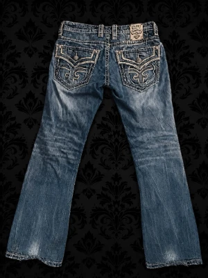 Rock Revival vintage jeans - Rock Revival jeans med slitningar och distressed detaljer framtill. Jeansen har raka ben med lite flare, broderade bakfickor med unika mönster. Klassisk femficksmodell och låg midja. Köptes i Köpenhamn för 2500kr