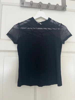 Svart mesh topp med kort ärm - Snygg svart topp med meshdetaljer över axlar och bröst. Kortärmad modell med hög hals och tight passform. Perfekt för dig som gillar en trendig och edgy stil.