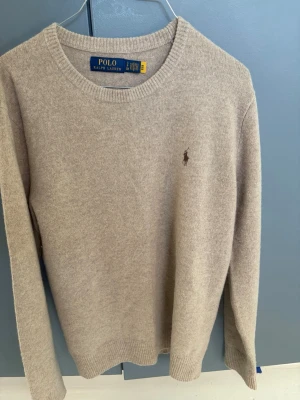 Beige stickad tröja Polo Ralph Lauren - Klassisk beige stickad tröja från Polo Ralph Lauren med rund halsringning och diskret broderad logga på bröstet. Tröjan är helt ny och aldrig använd. 