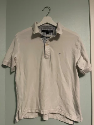 Vit pikétröja från Tommy Hilfiger - Klassisk vit pikétröja från Tommy Hilfiger med korta ärmar och krage. Tröjan har diskret logga på bröstet och randigt tyg på insidan av kragen. Perfekt för en clean och stilren look.