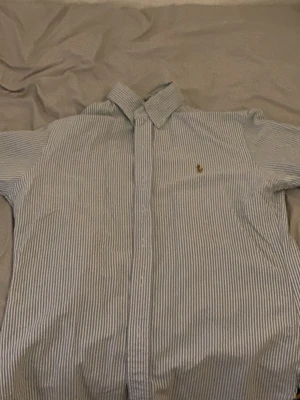 Randig skjorta från Polo Ralph Lauren - Klassisk skjorta från Polo Ralph Lauren i vitt och ljusblått randigt mönster. Skjortan har krage, långa ärmar och knappstängning framtill samt den ikoniska broderade loggan på bröstet. Perfekt för en clean och stilren look.