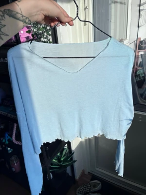 Ljusblå croppad långärmad topp - Trendig ljusblå croppad topp med lång ärm och rå kant nertill. Toppen har en v-ringad halsringning och är tillverkad i ett mjukt, ribbat material som ger en avslappnad vibe. Perfekt att styla med höga jeans eller kjol. Aldrig använd endast testad