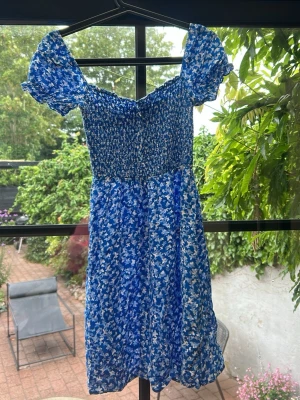 Blå blommig klänning från FB Sister - Söt blå klänning från FB Sister med vitt fjäril/blommönster och smockad överdel. Klänningen har korta puffärmar och är tillverkad i ett lätt och luftigt material, perfekt för varma dagar. Den har en somrig look med volangdetaljer vid ärmslut.