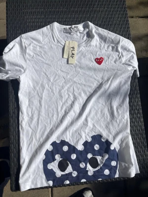 Vit t-shirt med hjärtdetalj och prickar - Vit t-shirt från Comme des Garçons PLAY med röd hjärtdetalj på bröstet och stort blått hjärta med vita prickar nedtill. Klassisk rund hals och korta ärmar. Perfekt för dig som gillar streetwear och lekfull design.
