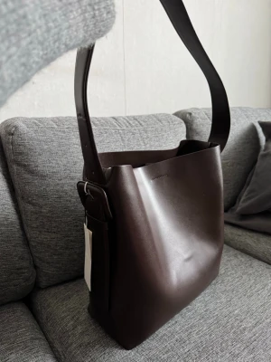 Mörkbrun totebag Arket - Snygg mörkbrun handväska i bucket-modell, tillverkad av 100% kromfritt garvat läder. Väskan har bred axelrem och minimalistisk design utan synliga loggor. Perfekt för dig som gillar stilrena accessoarer med modern känsla.
