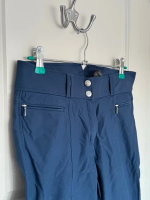 Mörkblå ridbyxor med dragkedjor - Snygga mörkblå ridbyxor med hög midja, två silverfärgade knappar och dragkedjefickor framtill. Byxorna har förstärkta partier på insidan av benen och stretchigt material för extra komfort vid ridning.