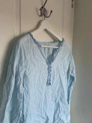 Ljusblå blus med volangkant - Söt ljusblå blus med långa ärmar och dekorativ volangkant längs halsringningen och knäppningen. Blusen har en avslappnad passform och är tillverkad i ett tunt, mjukt tyg. 