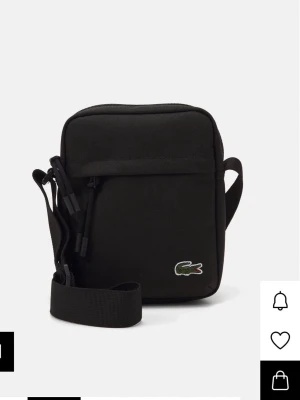 Svart axelväska från Lacoste - Snygg svart axelväska från Lacoste i kompakt modell med justerbar axelrem. Väskan har dragkedja upptill, en ficka framtill och den klassiska gröna krokodilloggan broderad på framsidan. Tillverkad i slitstark polyester.