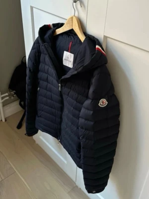 (Äkta) Mörkblå Moncler Eus Giubbotto Dunjacka - Otroligt snygg dunjacka från Moncler som är perfekt tjocklek nu till våren. Jackan är mörkblå och otroligt snygg med de klassiska stripesen på luvan. Väldigt fint skick. Storlek 2 som motsvarar M (passar även S). OBS! Jackan har en liten lagning vid loggan som syns på bild 3. Tveka inte på att meddela vid frågor.