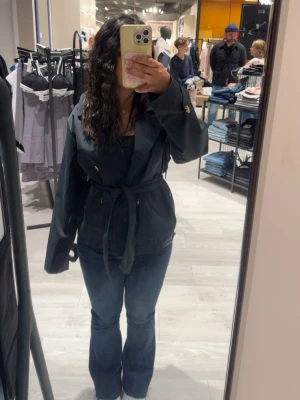 Mörkblå trenchcoat med bälte - Snygg mörkblå trenchcoat med knytbälte i midjan och långa ärmar. Jackan har klassisk krage och en loose passform som ger en avslappnad men ändå stilren look. Perfekt för dig som vill ha en enkel men trendig jacka till vardags.