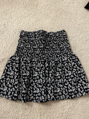 Svart blommig kortkjol från Gina Tricot - Supersöt svart kortkjol från Gina Tricot med vitt blommönster. Kjolen har smock i midjan för en bekväm passform och volang nedtill som ger en extra touch. Perfekt att styla med en enkel topp eller hoodie.