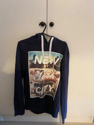 Mörkblå hoodie New York City Divided - Snygg mörkblå hoodie från H&M Divided med stort färgglatt New York City-tryck på bröstet och vita dragsnören i huvan. Tillverkad i mjukt bomullsmaterial och har långa ärmar. Perfekt för dig som gillar streetstyle och vill sticka ut.