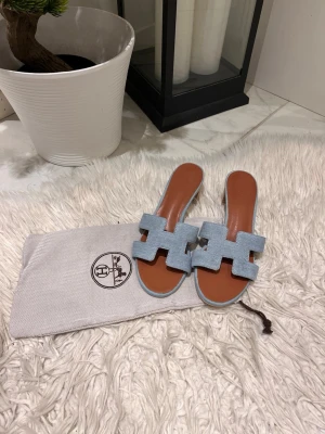 Blåa Oran sandaler  - Säljer ett par stilrena Oran sandaler i ljusblå denim med klassisk H-design. Innersulan är i brunt läder och ovandelen i mjukt tyg. Perfekta för en trendig och avslappnad look.