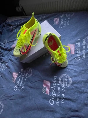 Nike Mercurial gul fotbollssko - Säljer ett par Nike Mercurial fotbollsskor i neon-gult med svarta och rosa detaljer. Skorna har snörning, låg profil och är tillverkade i ett lätt syntetmaterial för bra bollkontroll och grepp på planen. Perfekta för dig som vill sticka ut på fotbollsplanen.