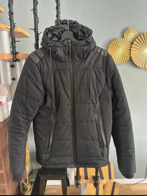 Svart vårjacka/höstjacka med huva storlek s - Säljer en svart pufferjacka med huva och dragsko. Jackan har två snedställda fickor med dragkedja framtill och justerbar nederkant. Quiltad design och diskreta reflexdetaljer på axlar och huva. Perfekt för kalla vinterdagar.