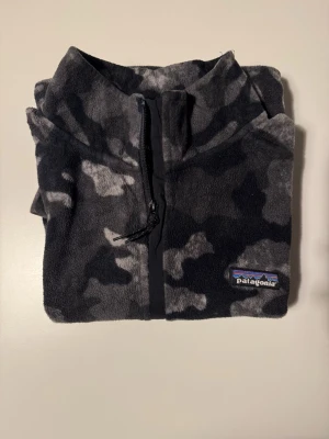 Patagonia, camo grå/svart, M, herr - Patagonia quarter-zip i mjuk mikrofleece med botaniskt camo-mönster i grått och svart. Stl muddar och nederkant, bröstlogga. Aldrig använd, utan lapp. Helt felfri. Stl M herr. Skicka mig gärna era erbjudanden