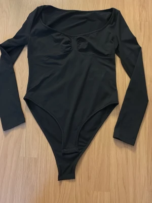 Svart långärmad body - Stilren svart body med långa ärmar och rundad urringning. Materialet är stretchigt och mjukt, vilket ger en bekväm passform. Bodyn har en lätt rynkning vid bysten för extra detalj. Perfekt att matcha med jeans eller kjol.
