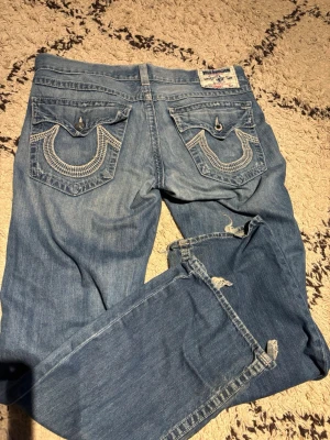 True Religion ljusblå jeans - Säljer ett par ljusblå jeans från True Religion med ikoniska broderade fickor bak och detaljerade sömmar. Klassisk rak passform och tillverkade i slitstarkt denim. Perfekta för dig som gillar streetwear och vill sticka ut med snygga detaljer.