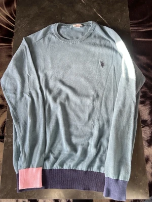 Blå tröja från U.S. Polo Assn. - Ljusblå tröja från U.S. Polo Assn. med rund halsringning och broderad logga på bröstet. Tröjan har ribbade muddar i mörkblått och en detalj i rosa på ena ärmen. Perfekt för dig som gillar stilrena plagg med en twist.