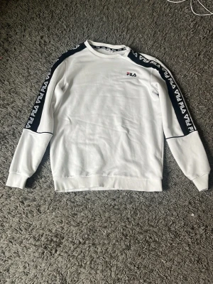Vit sweatshirt från FILA med logga - Snygg vit sweatshirt från FILA med svarta detaljer och logga på bröstet. Tröjan har svarta band med FILA-tryck längs ärmarna och ribbade muddar vid hals, ärmslut och nederkant. Perfekt för en sportig och avslappnad stil.