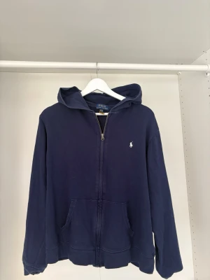 Mörkblå hoodie från Polo Ralph Lauren - Snygg mörkblå hoodie från Polo Ralph Lauren med dragkedja framtill och klassisk logga broderad på bröstet. Hoodien har huva, kängurufickor och ribbade muddar. Perfekt för en avslappnad och stilren look.