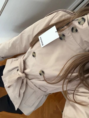 Beige trenchcoat från Bershka - Helt ny beige trenchcoat från Bershka med dubbelknäppning och stora knappar. Jackan har ett knytbälte i midjan och två fickor framtill. 