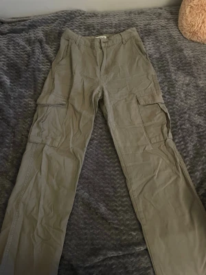 Gröna cargobyxor från Pull&Bear - Säljer ett par gröna cargobyxor från Pull&Bear med raka ben och stora sidofickor. Byxorna har klassisk cargomodell med knapp och dragkedja framtill. Tillverkade i ett mjukt bomullstyg som är skönt att bära.