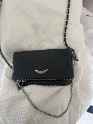 Mini Zadig Voltaire väska - Snygg svart axelväska i skinnimitation med silverfärgade detaljer och kedjerem. Väskan har ett emblem med vingar framtill och små nitar längs kanten. Perfekt storlek för det viktigaste och ger en cool vibe till din outfit.