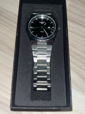Tissot PRX klocka med stållänk - Säljer en stilren Tissot PRX med svart urtavla och länk i rostfritt stål. Klockan har minimalistisk design, datumvisning och Tissot-logga på både urtavla och spänne. Snygg och tidlös accessoar som passar till allt.