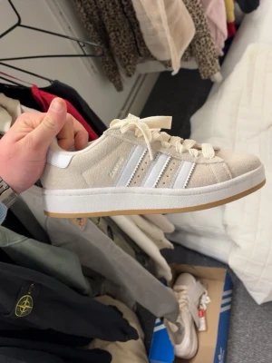 Beige Adidas sneakers med mocka - Snygga beige sneakers från Adidas i mocka med klassiska vita ränder på sidan och vit sula med gummidetalj. Skorna har lågt skaft och vita skosnören. Perfekta för en clean och stilren look. Helt nya och oanvända 