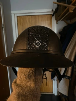 Svart glittrig ridhjälm Charles Owen - Snygg svart ridhjälm från Charles Owen med glittrig detalj längs mitten och klassisk form. Hjälmen har ett justerbart hakband och en diskret logga framtill. Perfekt för dig som vill sticka ut i stallet med stilren och säker look.