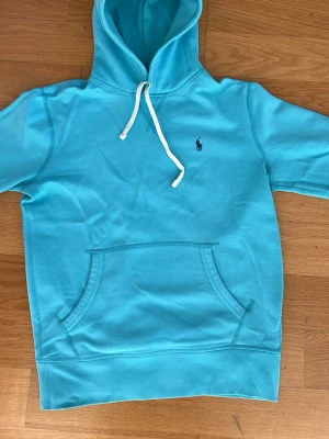 Turkos hoodie från Polo Ralph Lauren - Snygg turkos hoodie från Polo Ralph Lauren med vit dragsko i huvan och klassisk känguruficka framtill. Liten mörkblå logga broderad på bröstet. Perfekt för en chill och avslappnad stil. Tillverkad i mjukt bomullsmaterial. Använd ett par gånger men inget som märks när man bär hoodien.
