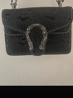 Svart axelväska med kedjerem - Snygg svart axelväska i ormmönstrat konstläder med silverfärgad kedjerem och ett coolt spänne i form av två drakhuvuden. Väskan har en kompakt, rektangulär form och detaljerade sömmar. Perfekt för dig som gillar edgy accessoarer.