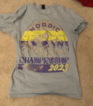 Nordic Championship 2023 T-shirt - Ljusgrå t-shirt med tryck i gult och lila som föreställer berg, skog och texten 'Nordic Championship 2023'. T-shirten har rund hals och korta ärmar, perfekt för dig som gillar grafiska motiv och sportiga vibbar.